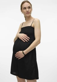 Robe de maternité noire avec un corsage froncé, des fines bretelles spaghetti et une jupe fluide. Le mannequin berce doucement son ventre, portant un bracelet simple.