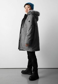 Dziecko stojące bokiem, ubrane w szary zimowy płaszcz z futrzanym kapturem, czarne spodnie, buty oraz niebieską wełnianą czapkę na tle jednolitym.