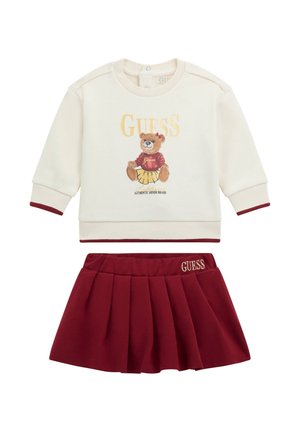 Maglione crema con accenti bordeaux, caratterizzato da una grafica di un orso e il testo "GUESS", abbinato a una gonna plissettata bordeaux con logo ricamato.
