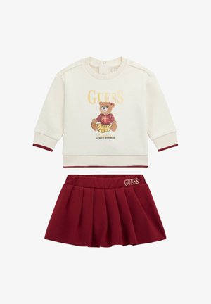 Cremfarbener Pullover mit bordeauxroten Akzenten, der eine Bären-Grafik und den Text "GUESS" zeigt, kombiniert mit einem plissierten bordeauxroten Rock mit gesticktem Logo.