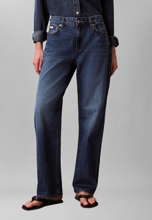 Personne portant un jean droit bleu foncé avec une chemise en denim rentrée et des tongs noires sur un fond neutre.