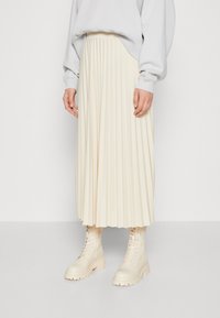 Falda midi plisada crema hecha de tela, combinada con botas de combate ligeras en color beis. El diseño presenta pliegues verticales y una textura suave.