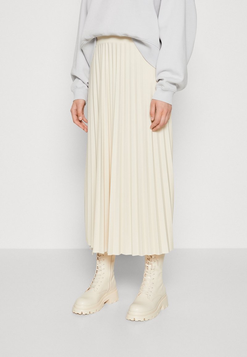 Falda midi plisada crema hecha de tela, combinada con botas de combate ligeras en color beis. El diseño presenta pliegues verticales y una textura suave.