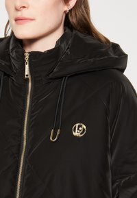 Giubbotto imbottito nero con zip frontale, colletto alto e cappuccio. Caratterizzato da un motivo a rombi e da un badge logo dorato sul petto.
