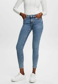 Jeans de mezclilla azul de tiro alto y ajustados, con cinco bolsillos, costuras sutiles y una clásica cremallera, combinados con zapatillas blancas.