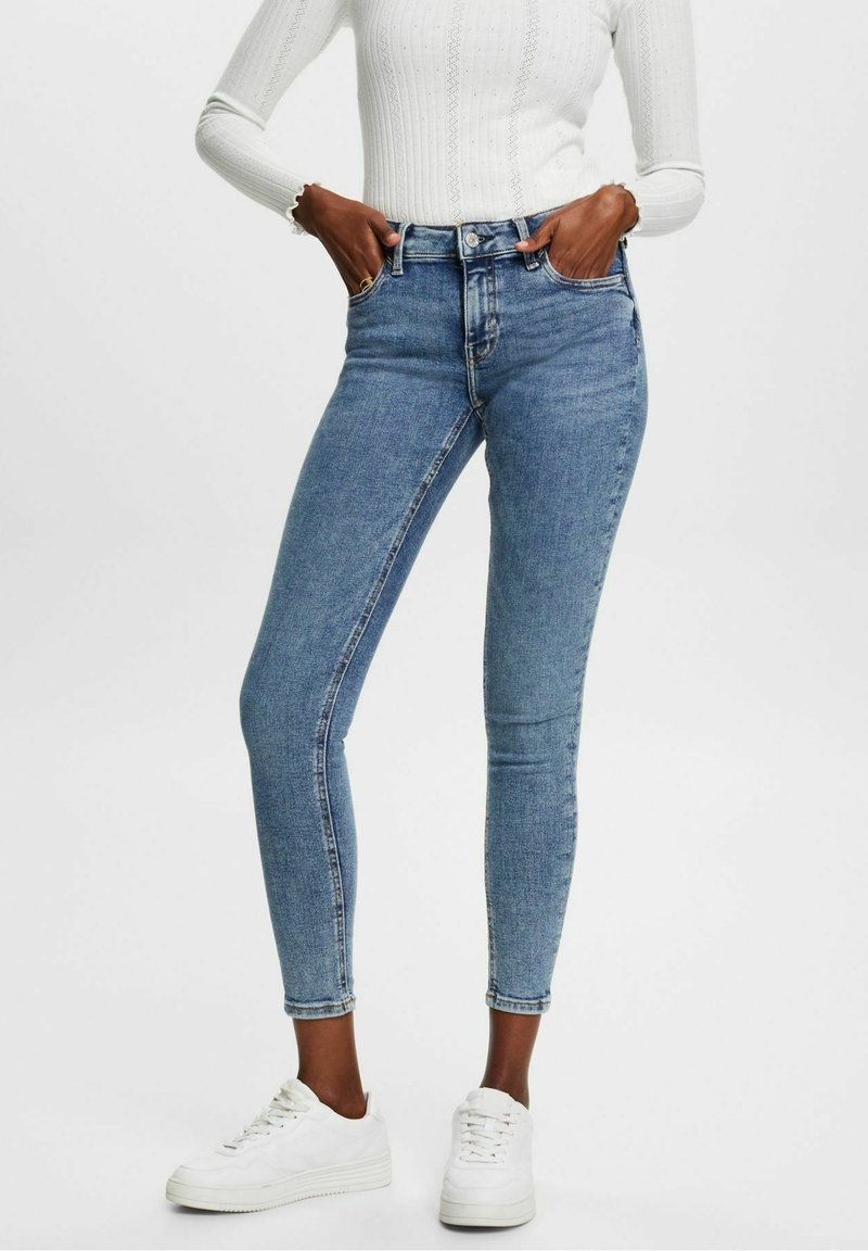 Jeans de mezclilla azul de tiro alto y ajustados, con cinco bolsillos, costuras sutiles y una clásica cremallera, combinados con zapatillas blancas.