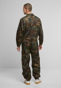 Zöld és barna árnyalatú camo overál, rugalmas derékkal és cipzáras zsebekkel; fehér cipővel párosítva.