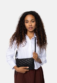 Donna con capelli ricci che indossa una camicia bianca e una gonna marrone tiene una borsa a tracolla in pelle intrecciata nera contro uno sfondo semplice.