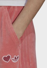 Pink velour sweatpants with a side pocket, featuring white embroidered heart and logo graphics, and ribbed waistband details.