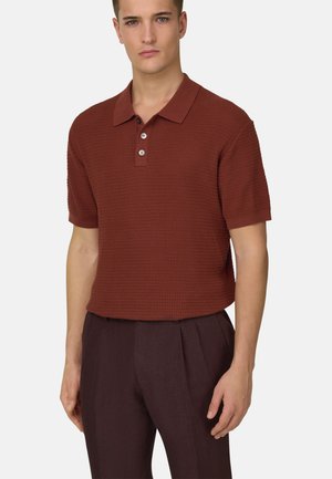 Ung mand iført strukturpræget rustbrun kortærmet polo med tre knapper og mørkebrune plisserede bukser, stående med neutralt udtryk.