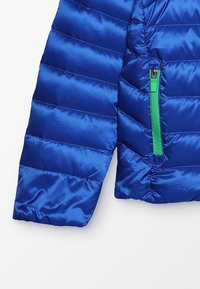 Modrá nafukovací bunda s lesklou, prošívanou texturou. Má zelenou kapsu na zip na boku. Lehký a izolační design.