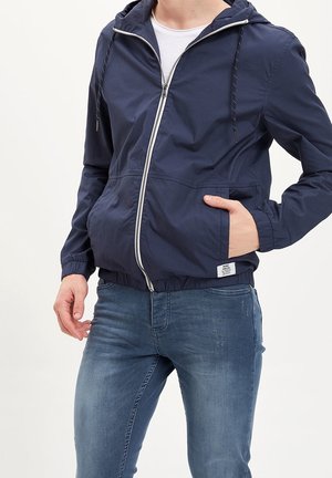 Mężczyzna noszący granatową kurtkę z kapturem na zamek z elastycznymi mankietami i ręką w kieszeni, w połączeniu z niebieskimi, przetartymi dżinsami.