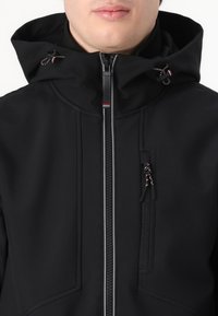 Casaco softshell preto com colarinho alto, fecho frontal de zíper e dois bolsos laterais. Apresenta capuz ajustável e uma costura decorativa subtil.