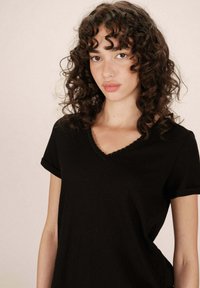 Giovane donna con capelli mossi e scuri, indossa una t-shirt nera a scollo a V, in piedi contro uno sfondo chiaro e uniforme, guardando leggermente di lato.