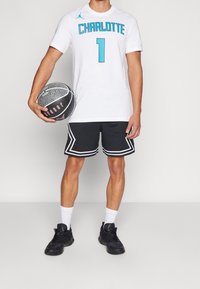 Camiseta blanca con estampado azul "CHARLOTTE 1", combinada con shorts de malla negros con ribete blanco. Sostiene un balón de baloncesto negro con superficie texturizada.