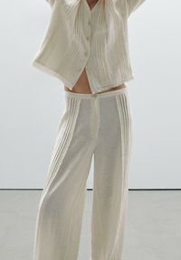 Lekki, teksturowany zestaw kremowy składający się z koszuli na guziki z pionowymi paskami oraz luźnych spodni o podobnych detalach i postrzępionych brzegach.