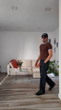 Muškarac u naočalama za sunce, s kapom, smešom košuljom i crnim hlačama prolazi pored bež sofe sa staklenim stolićem za kafu na kojem se nalaze cveće u modernoj dnevnoj sobi.
