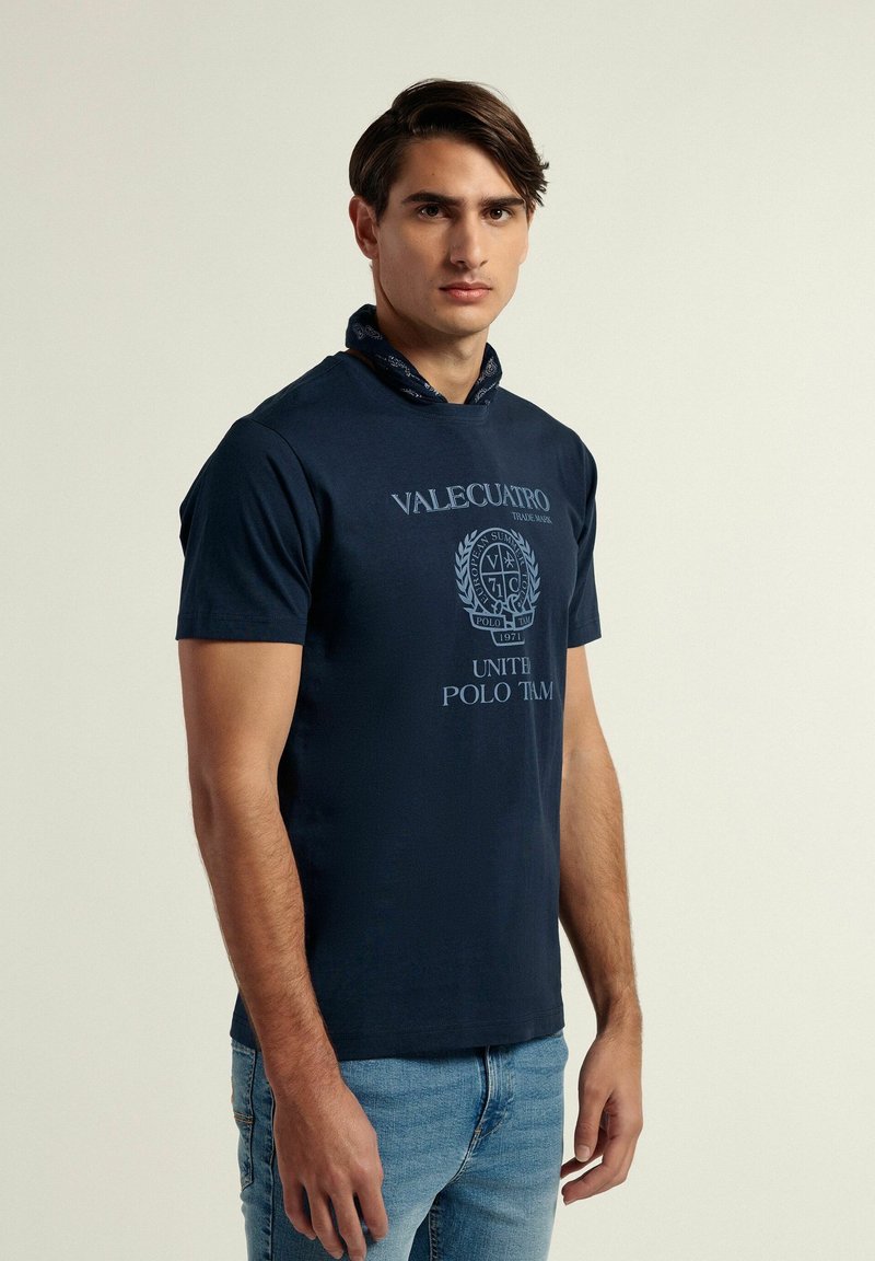 Camiseta de algodón azul marino con mangas cortas, con un diseño de logotipo y texto impreso en la parte delantera. Combinada con unos vaqueros azul claro.