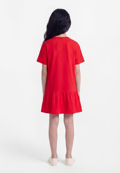 Otrok z dolgimi temnimi lasmi, oblečen v rdečo kratkoročno obleko z nabrano spodnjo obrobo, stoji obrnjeni stran na enobarvnem belem ozadju.