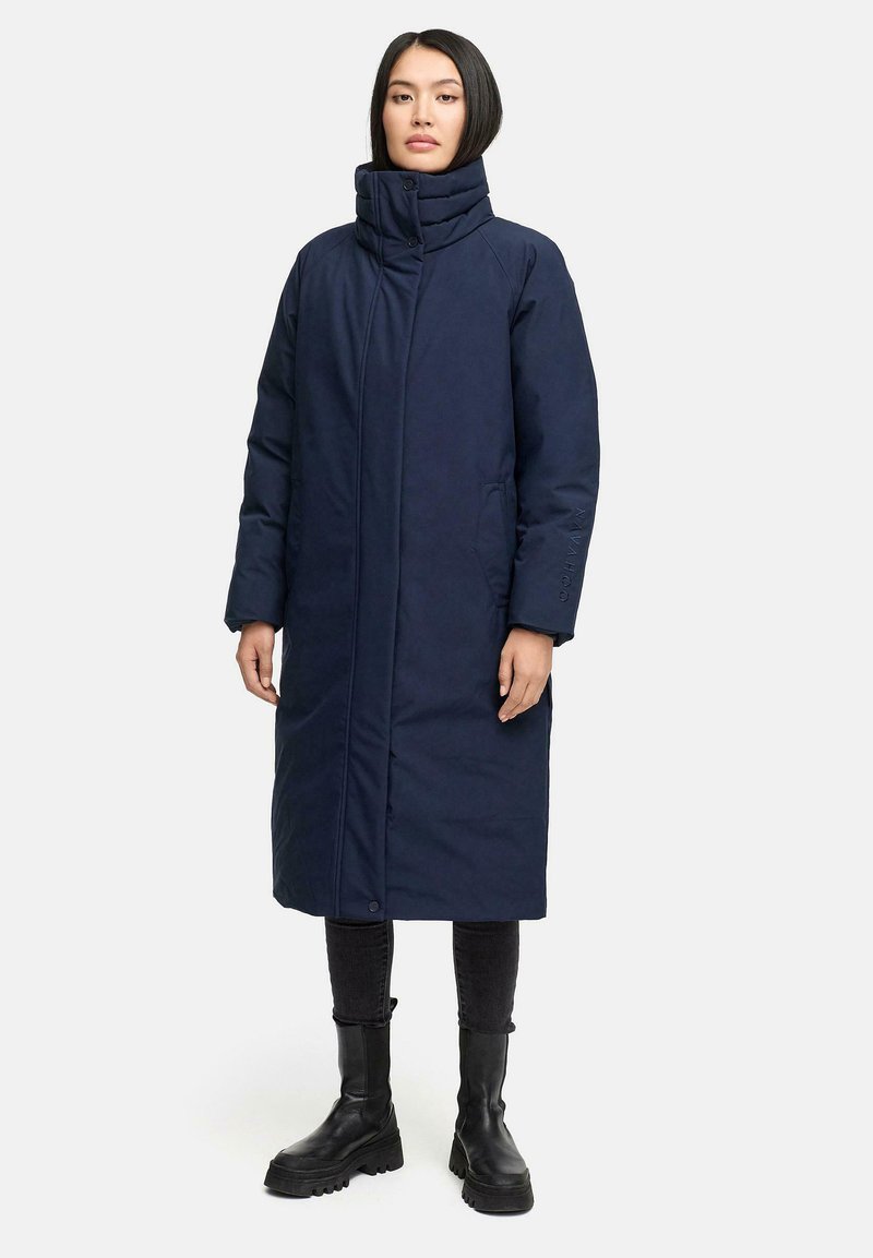 Marineblå polstret frakke med høj krave, hel længde design, to sidelommer og runde knapper, parret med sorte slim-fit bukser og støvler.