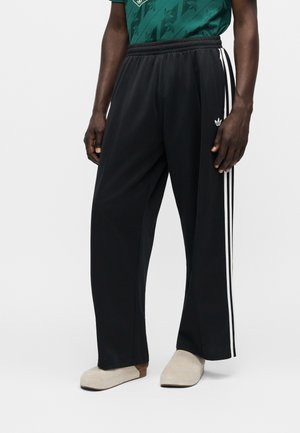 Mężczyzna noszący czarne spodnie dresowe Adidas z białymi paskami i logo, w połączeniu z beżowymi butami typu slip-on i zieloną koszulą w wzory.