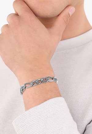 Pulsera de cadena de metal plateado en una muñeca, con eslabones entrelazados y un acabado pulido. Lucida con un suéter texturizado ligero.
