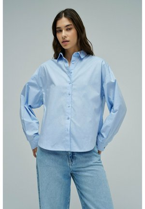 Femme portant une chemise bleu clair ample boutonnée et un jean bleu taille haute, debout devant un fond gris uni.