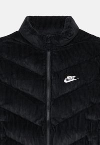 Juodas kordūrinis pūkinis švarkas su aukšta apykakle, užtrauktuku priekyje ir smailiais siūlais. Ant krūtinės puikuojasi balta Nike логотип.