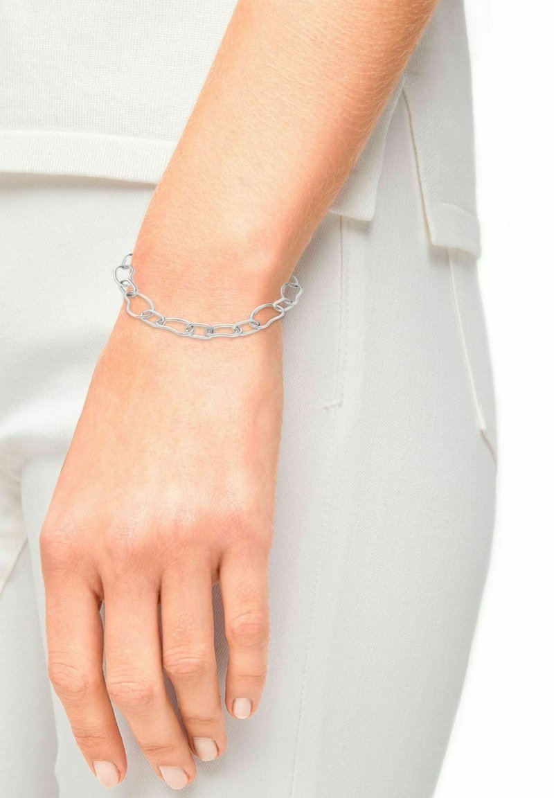 Main portant un bracelet à maillons en argent reposant à côté de la hanche, la personne vêtue d'un pantalon blanc et d'un haut blanc.