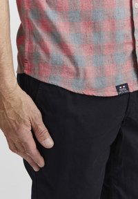 Camicetta a maniche corte a quadri in cotone rosso e grigio, con orlo arrotondato, abbinate a pantaloni neri con un'etichetta di tessuto blu. Mano appoggiata nelle vicinanze.