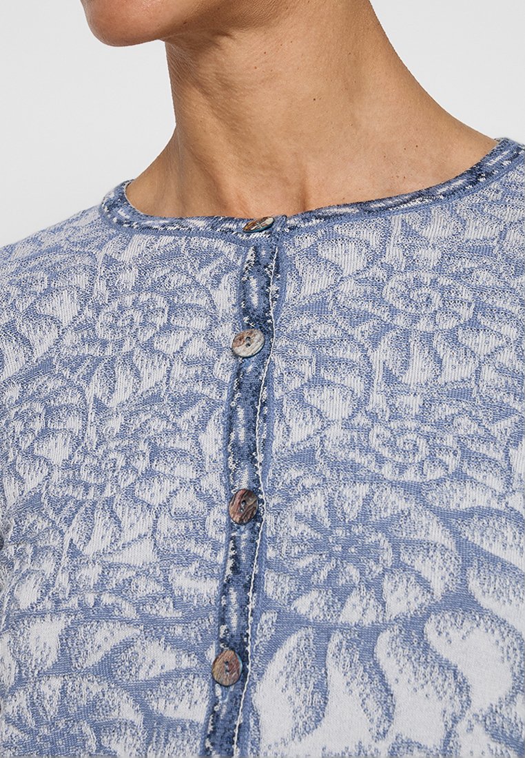 Gilet en tricot de couleur bleu clair et blanc avec un motif floral texturé. Il dispose d'un col rond et de six boutons en bois sur le devant.