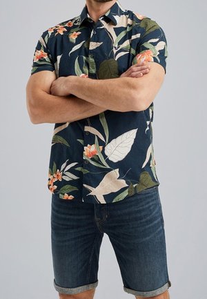 Homme les bras croisés portant une chemise boutonnée à manches courtes à motif floral sombre et un short en jean foncé retroussé, sur un fond uni.