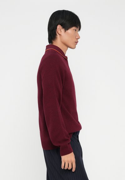 Pullover bordeaux con una finitura testurizzata, caratterizzato da un colletto a strisce arancioni in contrasto. Maniche corte e vestibilità rilassata, indossato con pantaloni a righe scure.