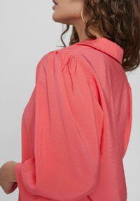 Blusa di colore corallo con una finitura testurizzata, dotata di maniche a palloncino e colletto. Il tessuto sembra leggero e leggermente lucido.