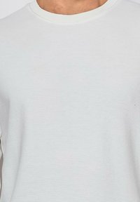 Chemise blanche à manches longues côtelées avec un col rond. Le tissu semble doux avec une surface texturée et des détails de couture minimaux.