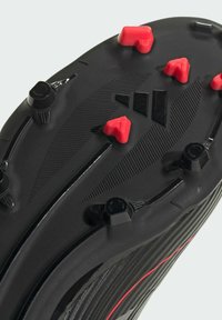 Semelle de crampons de football noirs avec des crampons rouges et noirs, surface texturée, rainures diagonales et un détail de logo central.