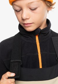Svart fleecejacka med en orange dragkedja och svarta remmar, hålls av ett barn. Designen har ett texturerat tyg och kontrasterande färger.