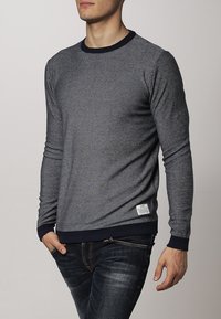 Donkerblauwe trui met lange mouwen en een gestructureerd grijs patroon, ronde hals en ribgebreide manchetten. Etiket op de onderste zoom voegt detail toe. G gedragen met donkere jeans.