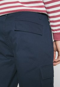 Osoba v námořnických modrých cargo kalhotách s kapsami na klopě a tričku s dlouhým rukávem v červenobílém pruhovaném vzoru.