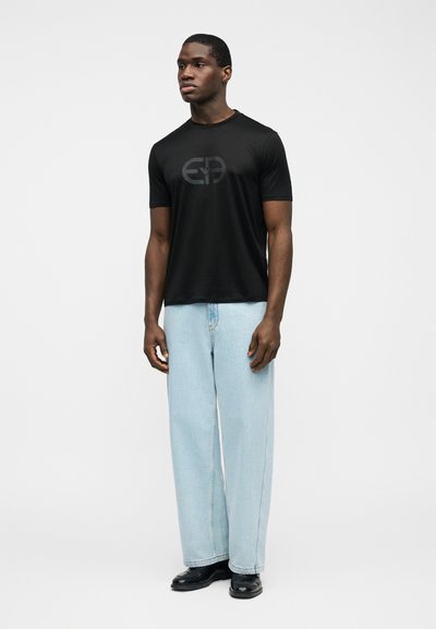 Άνδρας που στέκεται φορώντας μαύρο t-shirt με λογότυπο, ανοιχτό μπλε τζιν παντελόνι με φαρδιά μπατζάκια και μαύρα επίσημα παπούτσια, σε λευκό φόντο.