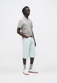 Sivá polokošeľa s malým vyšívaným logom, v kombinácii s svetlomodrými šortkami po kolená, sivými ponožkami a bielymi teniskami s hnedými podrážkami.