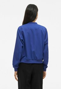 Femme aux longs cheveux foncés portant une veste bomber bleu royal et un pantalon noir, debout de dos contre un fond blanc uni.