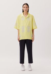 Camicia a maniche corte di colore giallo pallido con motivi geometrici bianchi e grigi. Abbinata a pantaloni a cropped blu navy e scarpe da ginnastica bianche.