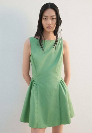 Vrouw met lang zwart haar, draagt een mouwloze groene jurk met een aangesloten taille en een uitlopende rok, staand tegen een effen witte achtergrond.