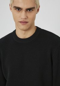 Pull en tricot côtelé noir avec un col rond et une finition texturée. Le tissu semble doux et ajusté, mettant en valeur le décolleté.