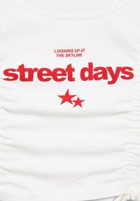 Bela majica brez rokavov z rdečim napisom "LOOKING UP AT THE SKYLINE" nad velikim napisom "street days" in dvema rdečima zvezdama pod napisom.