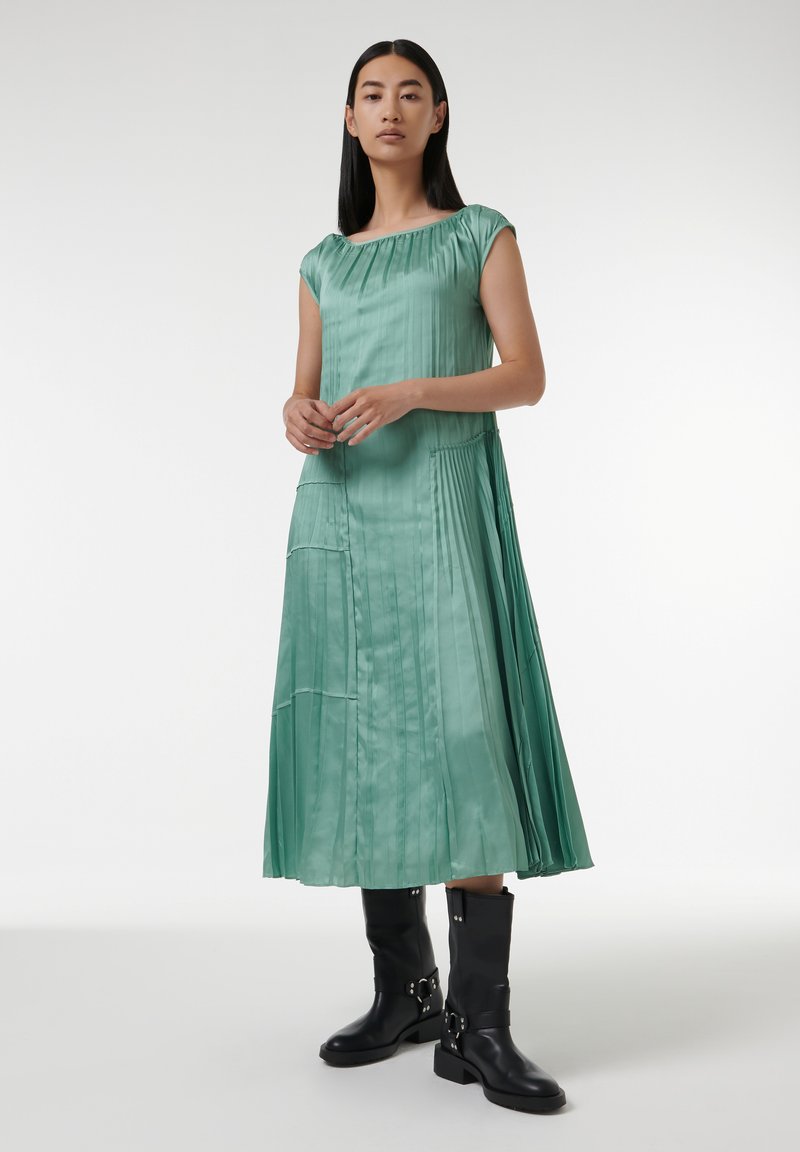 Mintgrünes, plissiertes Kleid mit Rundhalsausschnitt und kurzen Ärmeln, verziert mit vertikalen Streifen. Kombiniert mit schwarzen Kniefallstiefeln.