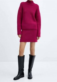 Fuksjowy sweter z dzianiny o wysokim dekolcie w zestawie z dopasowaną spódnicą dzianinową. Modelka nosi czarne skórzane kozaki za kolano.