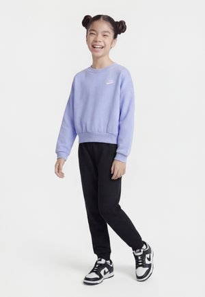 Šypsosi mergina su „space buns“ šukuosena, vilkinti levandų spalvos „Nike“ džemperį, juodus sportinius kelnes ir juodai baltus „Nike“ sportbačius, stovinti ant baltos fono.