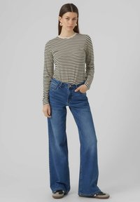 Gestreiftes Langarmshirt in Creme und Schwarz, kombiniert mit hochgeschnittenen, blauen Denim-Jeans im weiten Schnitt. Das Model trägt lässige Sneakers.